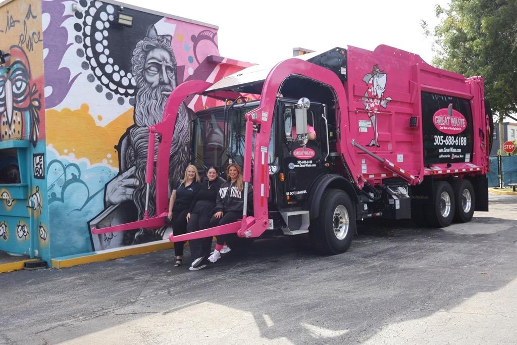 Angela, Amanda and Amber Piccinonna sitting together on the lowered front-loader arms of a pink Great Waste garbage truck carrying the Paulie the Great White Shark mascot, in front of a colorful painted mural