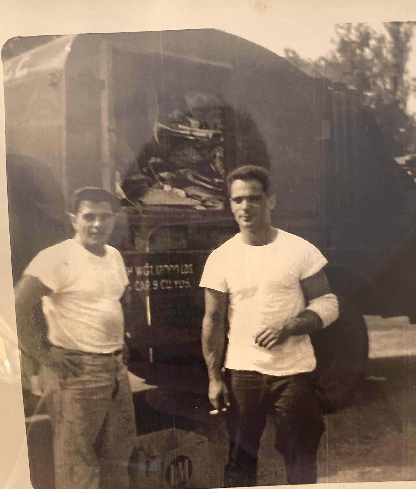 A weathered black-and-white photograph of two young men in white t-shirts standing shoulder to shoulder in front of a loaded rear-loading garbage truck